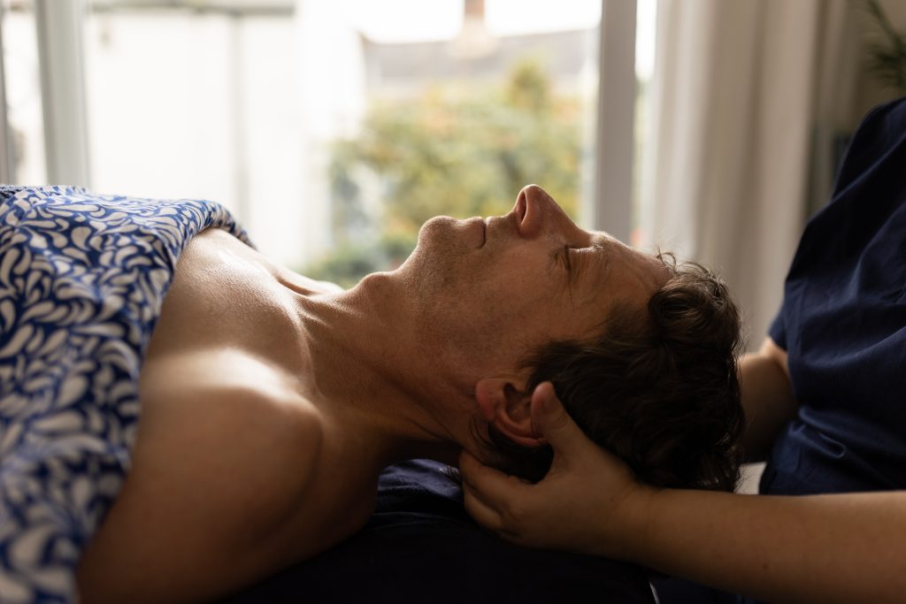 Person lying comfortably under a patterned blanket while a practitioner gently supports their head and neck during a relaxing myofascial release treatment.